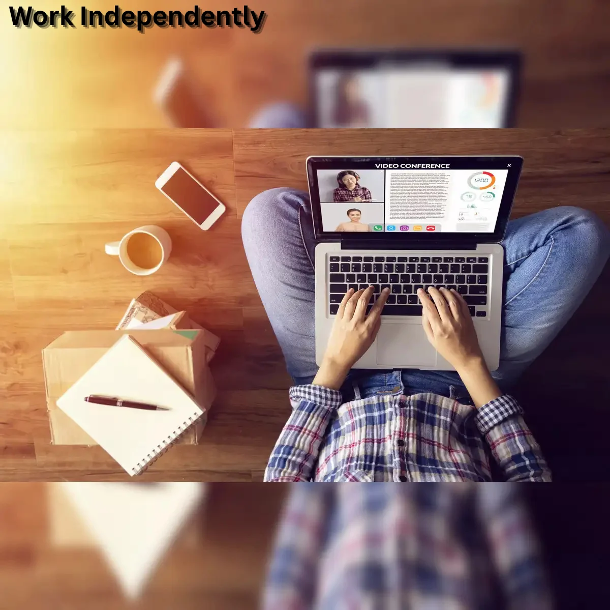 A person sitting on the floor with a laptop on their lap, working independently, with a notebook, phone, and coffee cup placed beside them on a wooden surface.